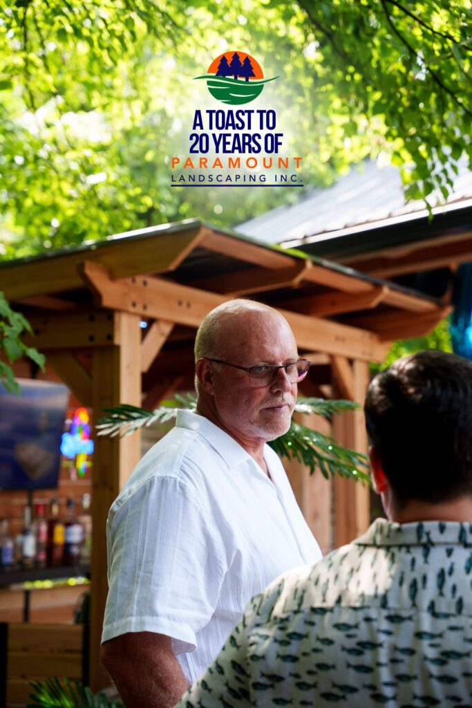 Paramount Landscaping President Jamie Wad stands under a wooden pergola at the 20th Anniversary event, with the branded balloon sign in the background.